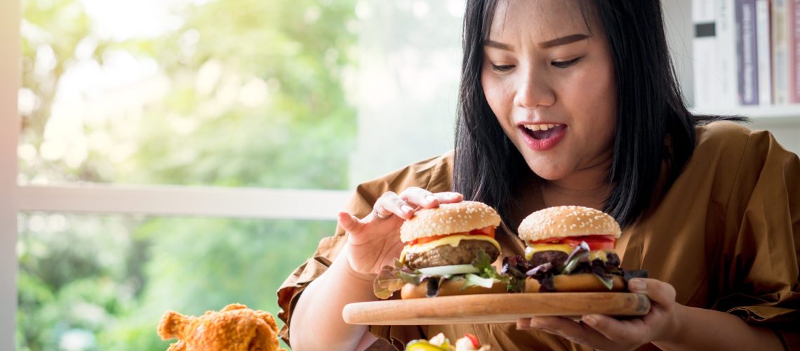 Hungry overweight woman holding hamburger on wooden plate after delivery man delivers foods at home. Concept of binge eating disorder (BED) and Relaxing with Eating junk food.